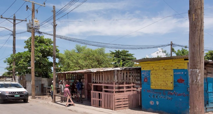 A small group of people in a poor village congregate casually outside a local shop under electricity distribution poles and a mass of associated wires.