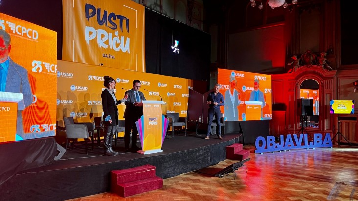 A man and a woman stand on a stage talking with another man in the background.