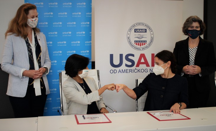 Two women at a table bump fists while two women stand behind them observing.