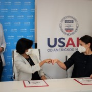 Two women at a table bump fists while two women stand behind them observing.